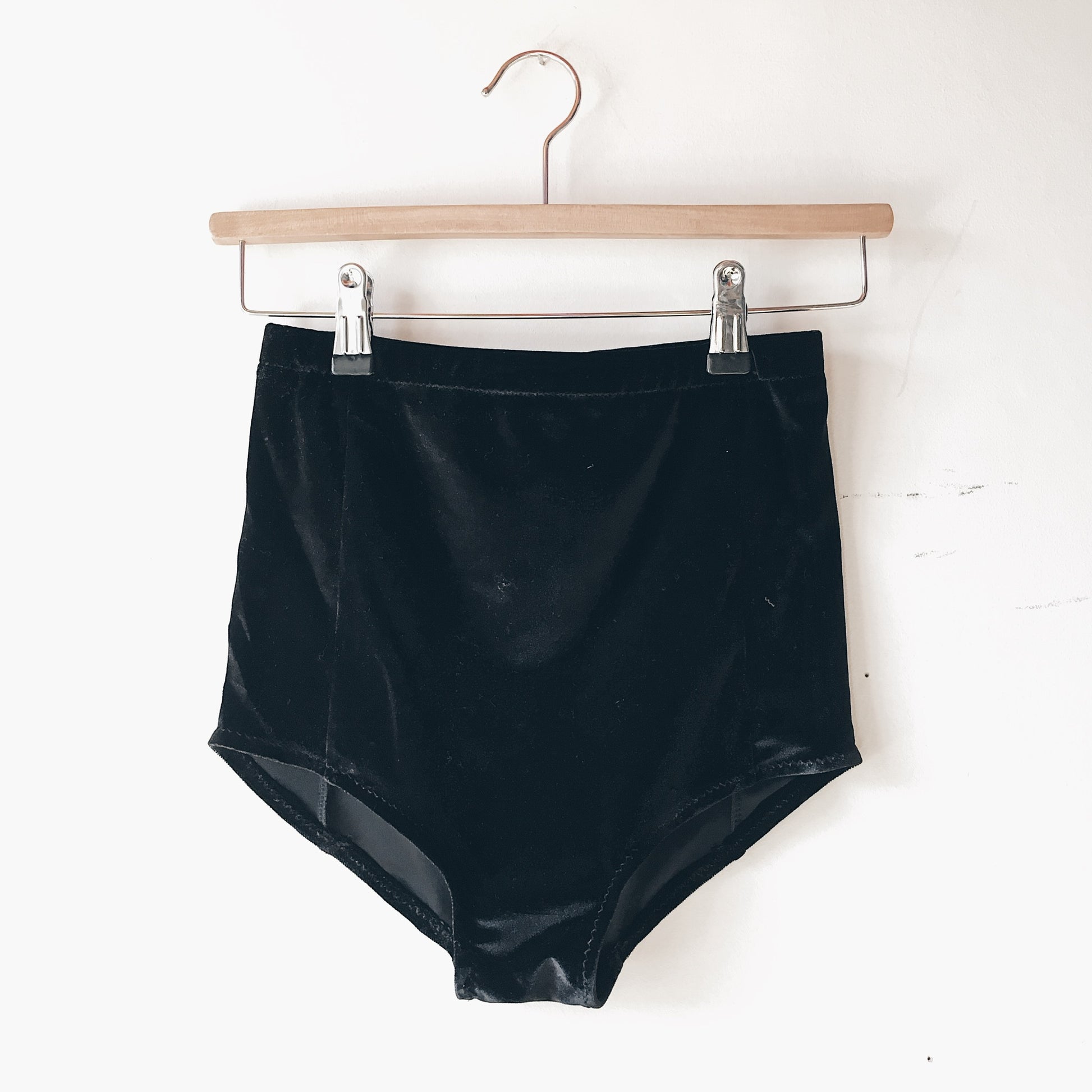 Black velvet booty shorts on a wooden hanger against a white wall