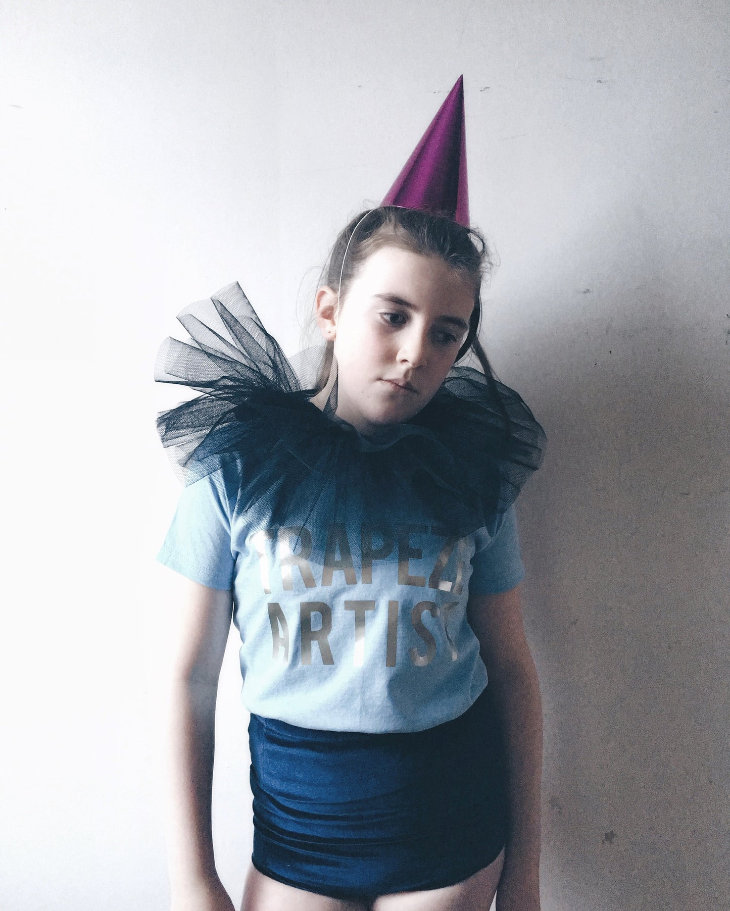 A girl in a 'circus' costume wearing a pink cone hat, black tulle collar, light blue trapeze artist t-shirt and blue velvet shorts