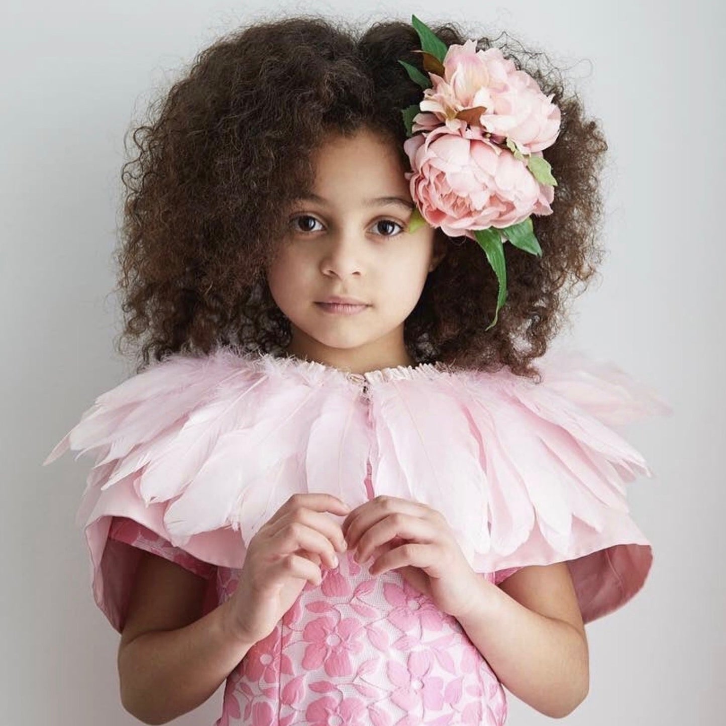 Girl wearing a pink floral brocade dress, pink feather cape, and large pink peonies in her brown curly hair