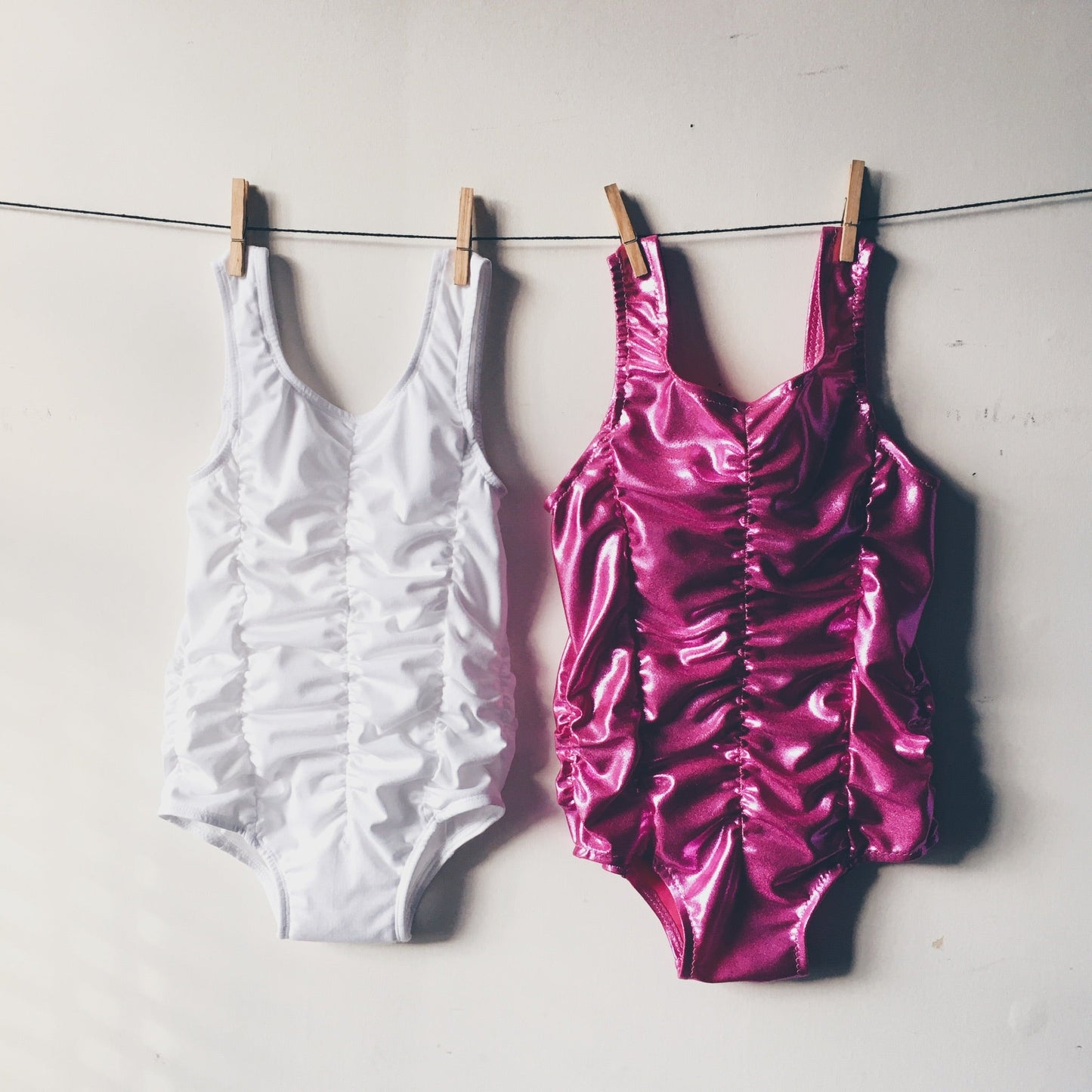 Two, Tallulah Ruched Leotards pegged on a line against a wall. White on the left and Pink on the right