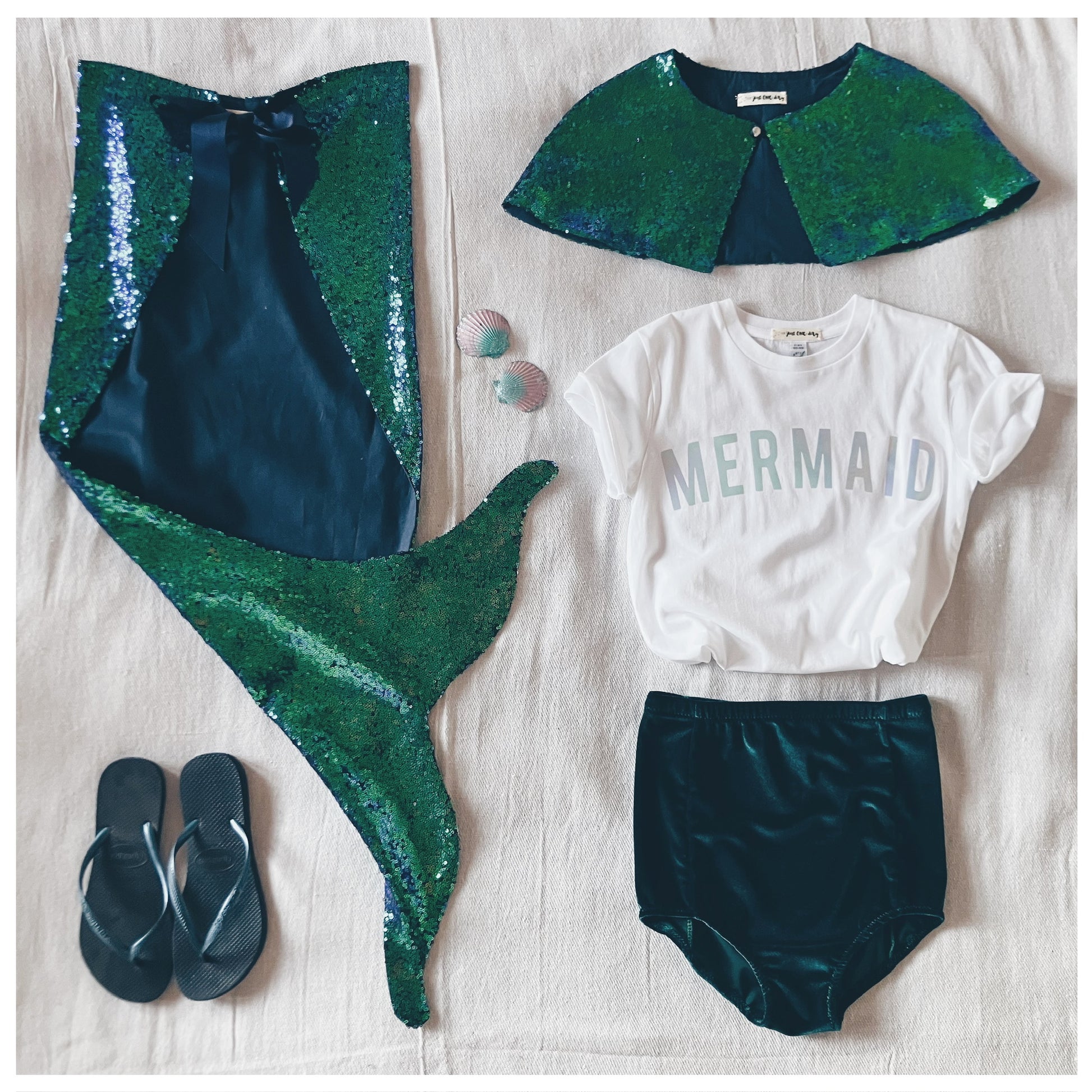 flat-lay of child's mermaid costume by For Just ONE Day - sequin cape, mermaid t-shirt, velvet shorts, 2 shell hair clips, mermaid tail, and some flip flops