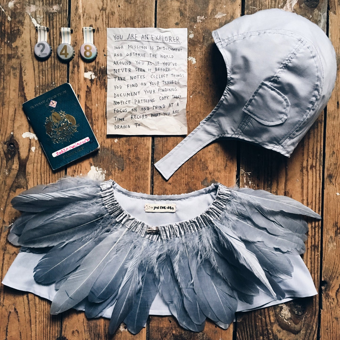 flat lay of a child's explorer costume, grey flying hat, feather cape, passport, explorer note and 3 medals. On wooden floorboards