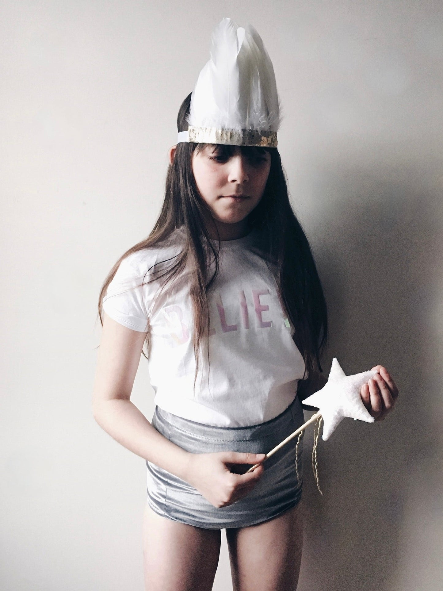 Child with long dark hair holding a star wand.  Wearing a white Believe t-shirt, feather headdress and grey shorts by For Just ONE Day
