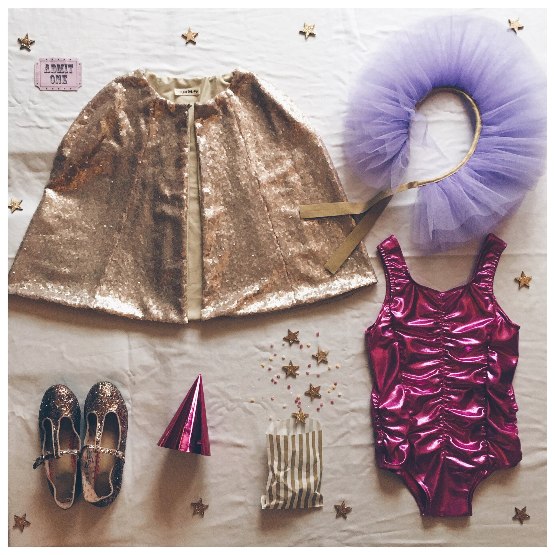 Flat lay of a child's circus costume - Gold Sequin Cape, Lilac Tulle Collar, Magenta Tullulah Leotard, on white background with scattered gold glitter stars
