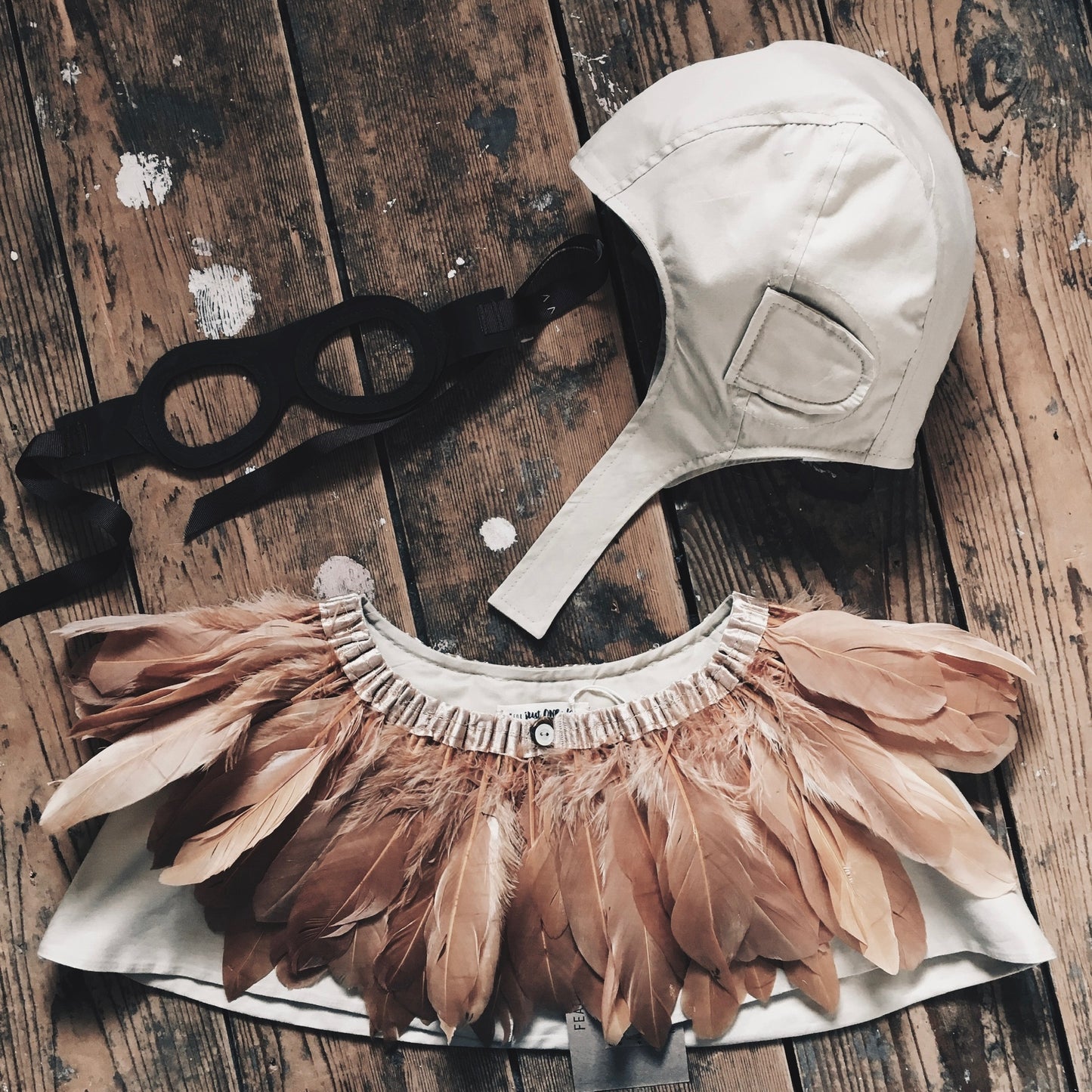 flat lay - a child's explorer costume arranged on wooden floorboards. Beige flying hat, copper feather cape and leather flying goggles