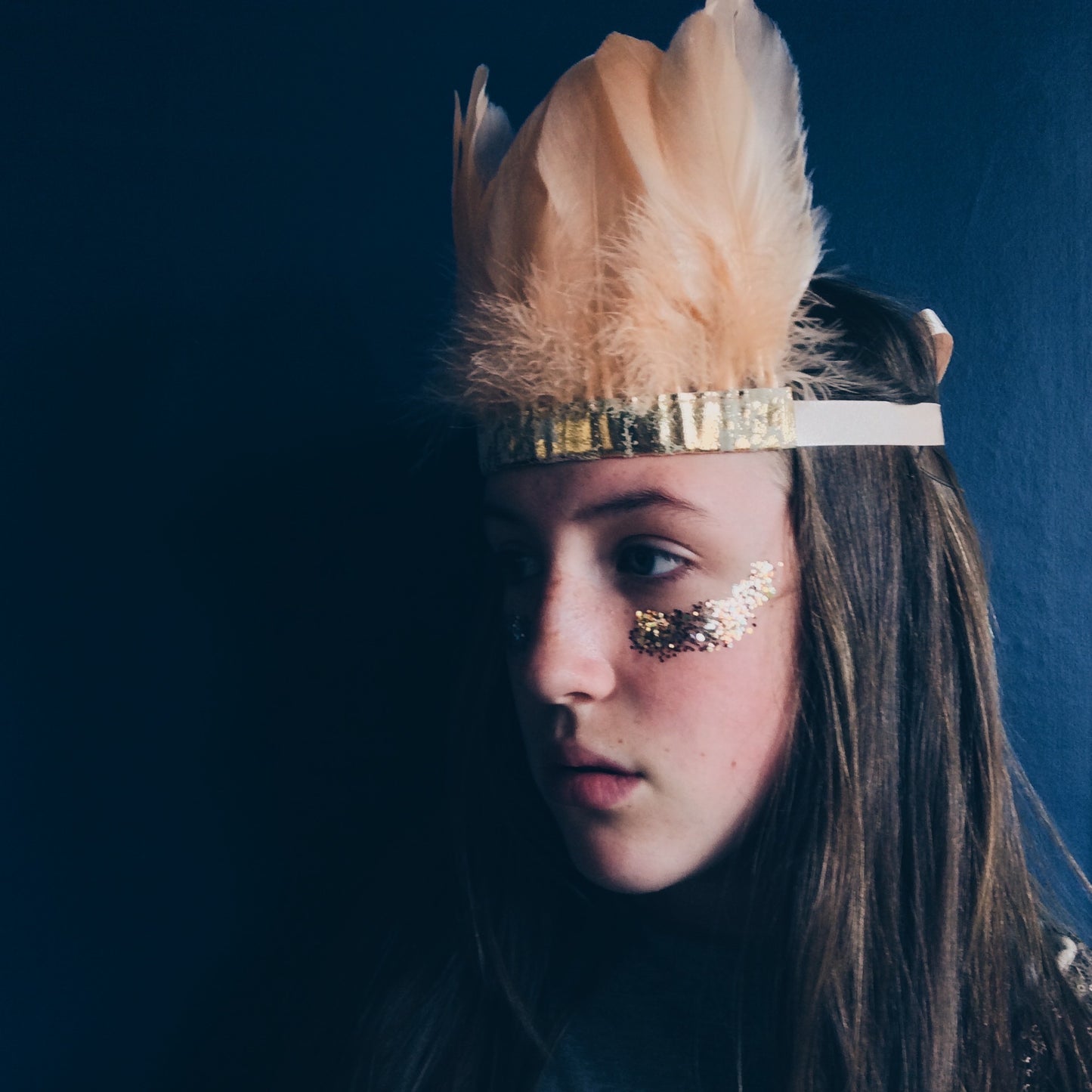 Girl with gold glitter on her cheeks against a dark blue wall.  Wearing a peach feather and metallic leather headdress by For Just ONE Day.