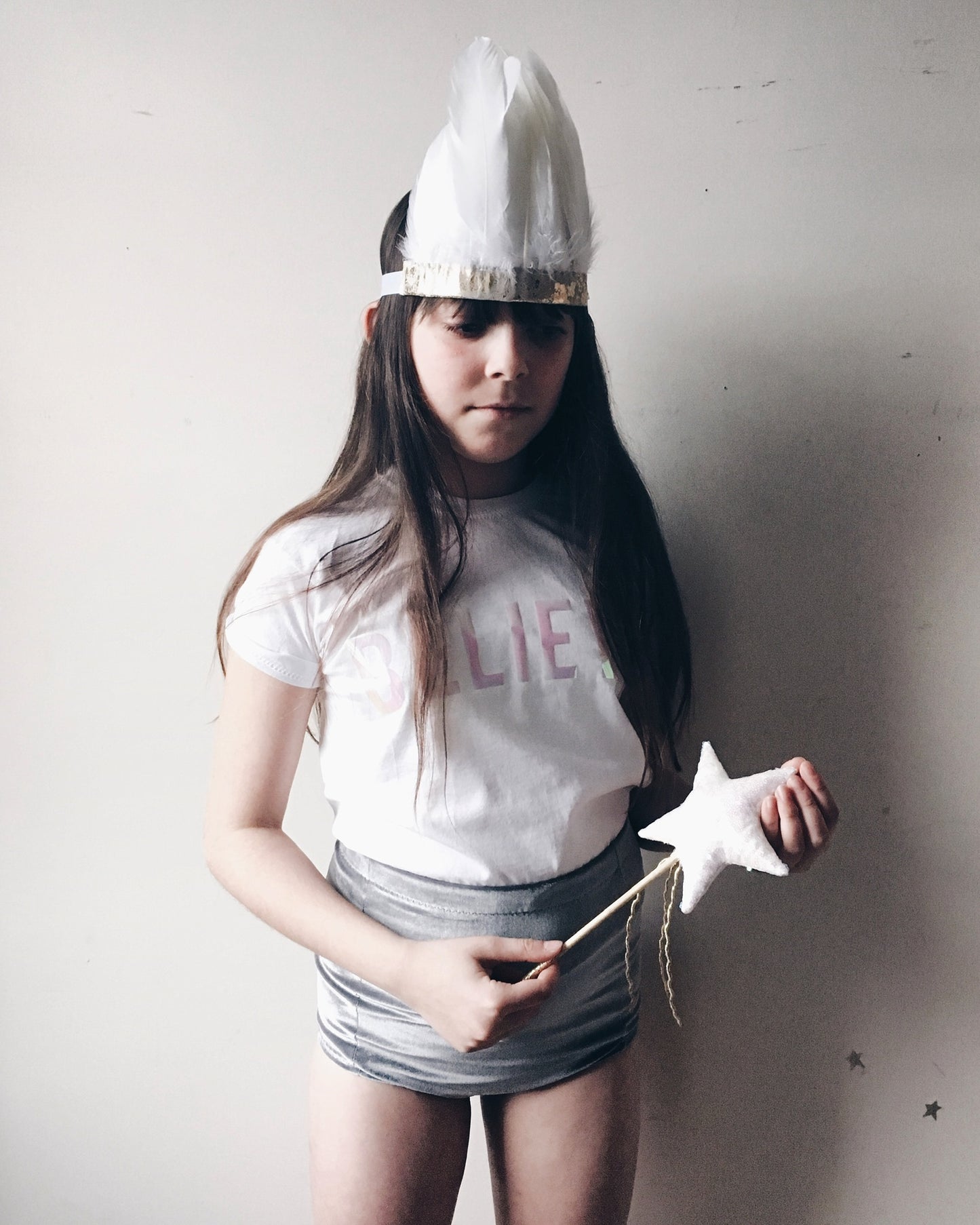 Girl stood against a white wall, holding a star wand and wearing a white feather headdress, believe t-shirt and grey velvet shorts
