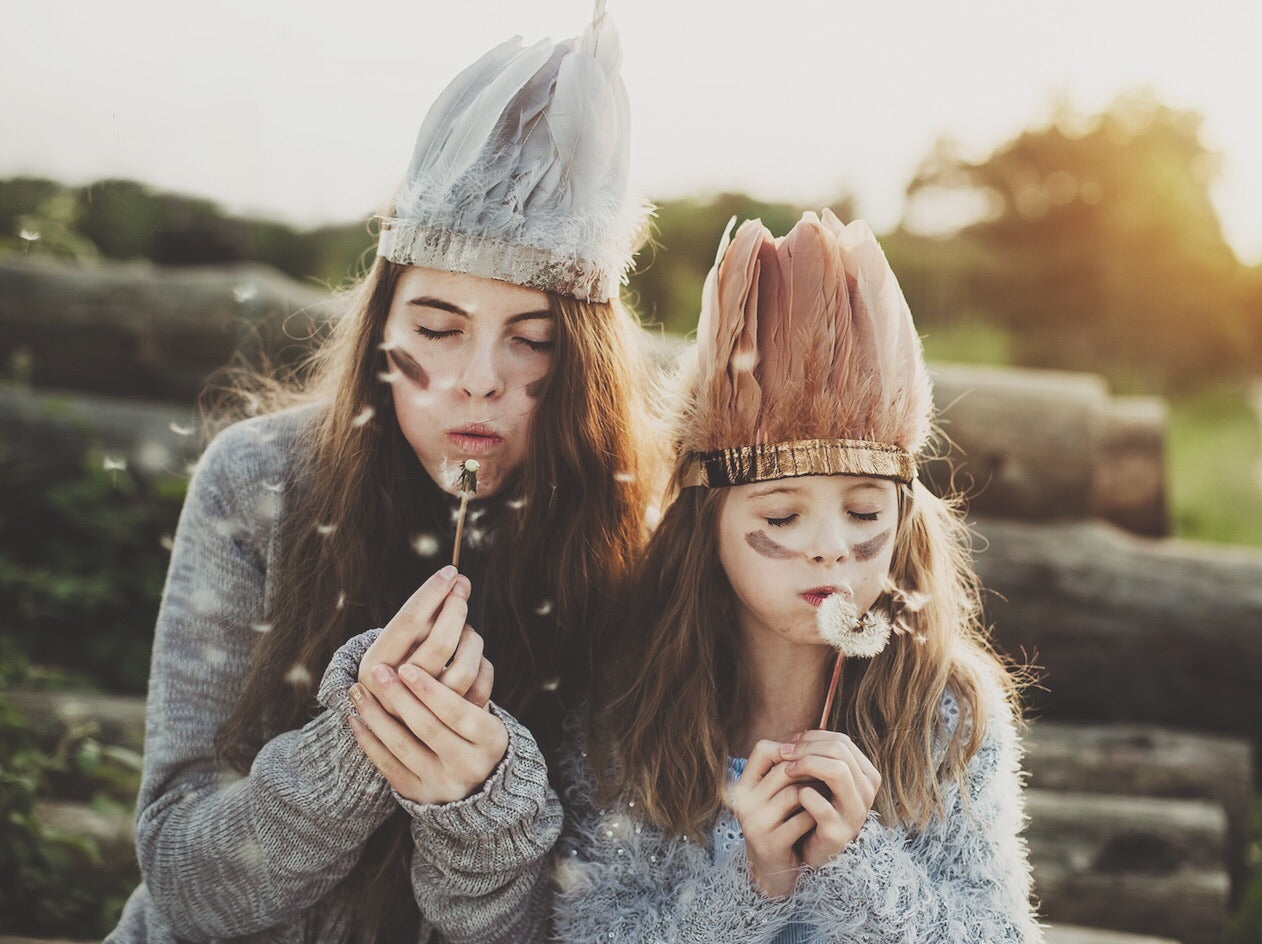 Two girls wearing grey, and copper feather headdresses, blowing dandelions