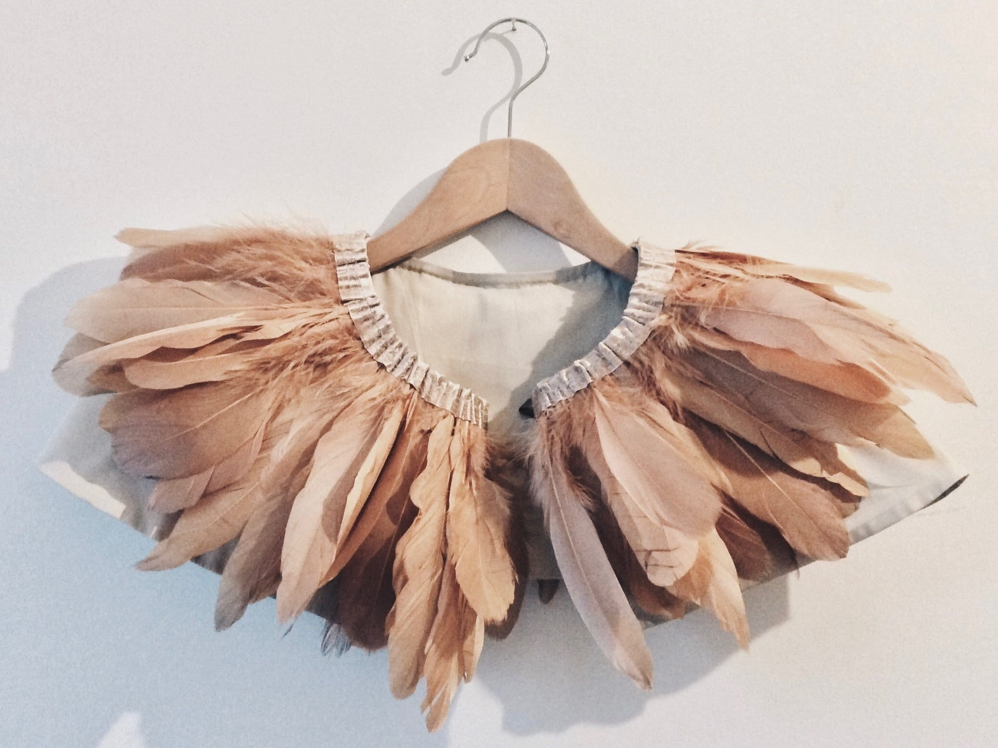 Copper coloured feather cape on wooden hanger against a white wall