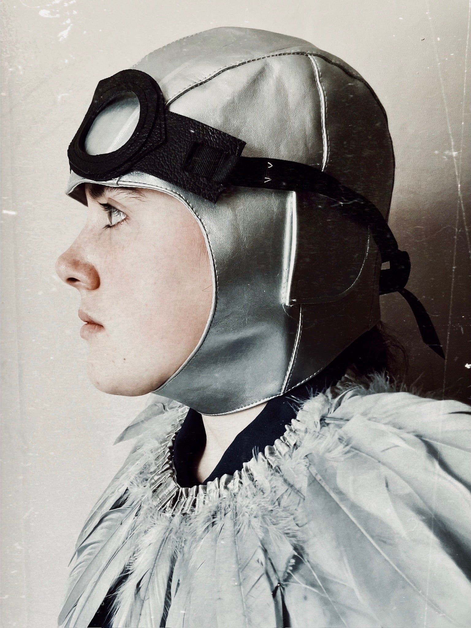 Child wearing a grey flying hat, leather goggles and a feather cape looking to the left
