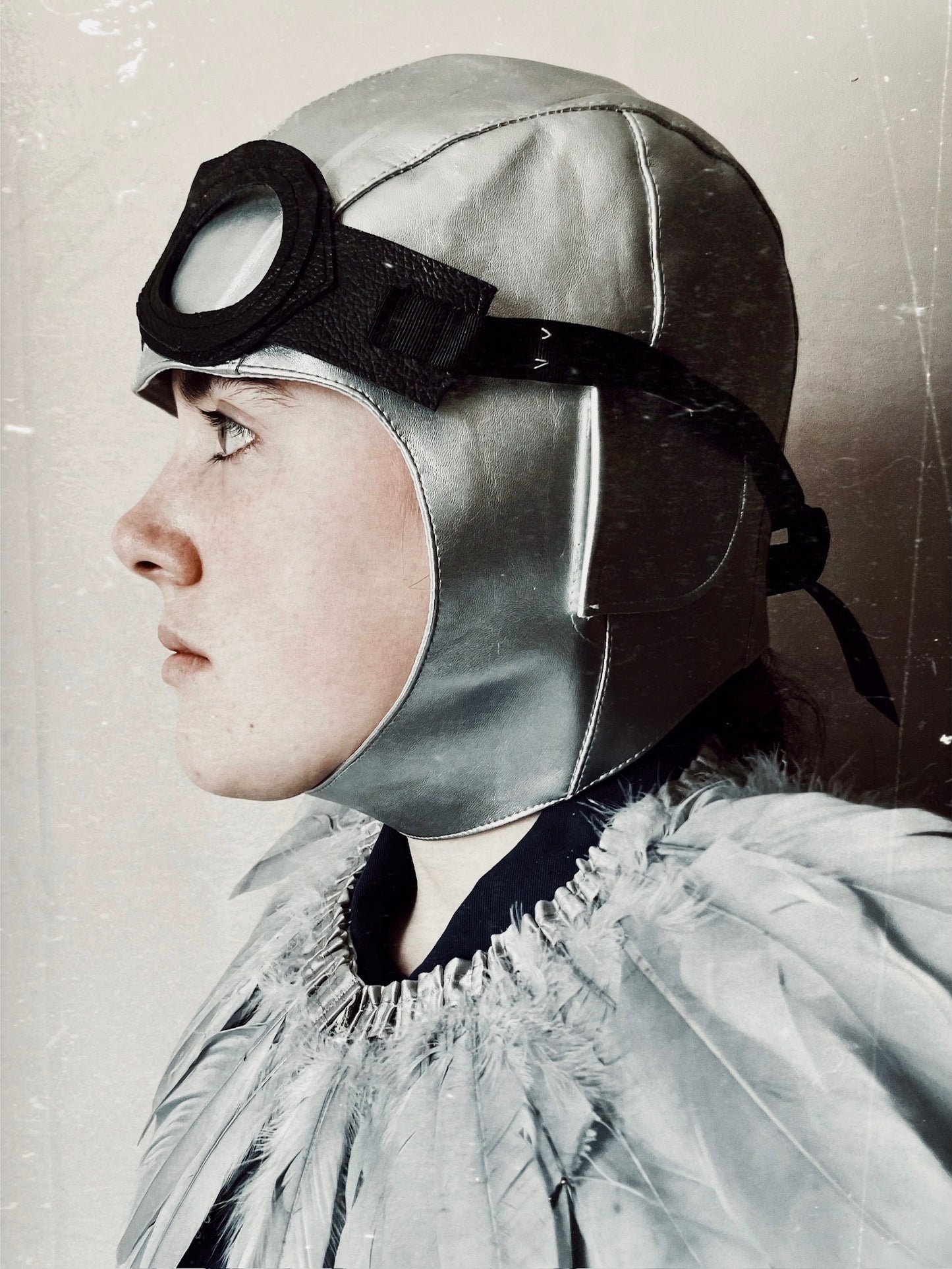 side view of a child wearing a grey vintage flying hat, a grey feather cape and leather flying goggles