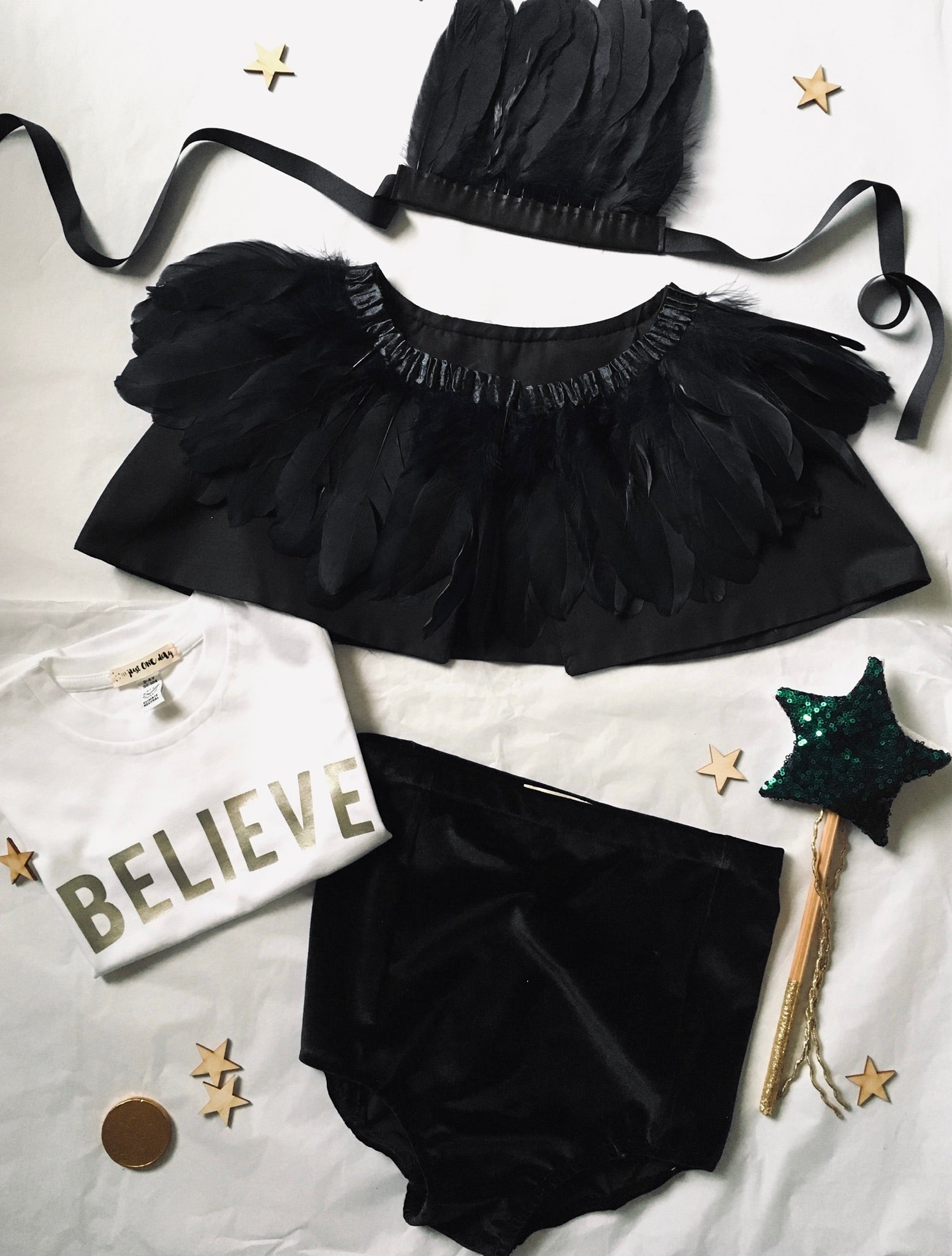 flat lay of black feather headdress, black feather cape, black velvet shorts and a believe t-shirt